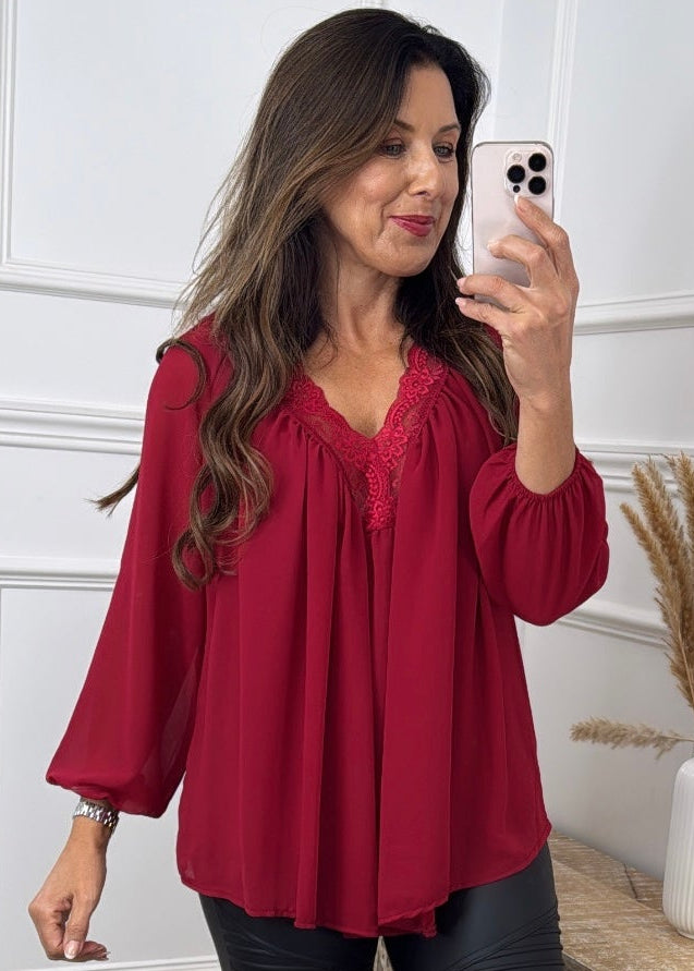 A woman takes a mirror selfie wearing a long-sleeve burgundy blouse with a tie-neck detail, paired with black leggings and black pointed-toe heels, in a decorated room with white paneled walls and a rustic wooden side table.
